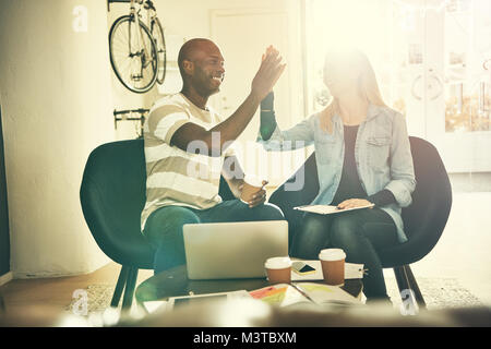 Two diverse young work colleagues smiling and high fiving each other while sitting in a stylish modern office Stock Photo