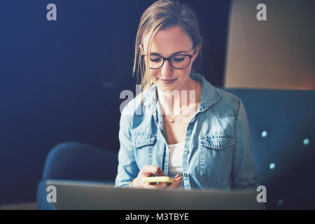 Smiling young businesswoman sitting on a sofa checking text messages on her cellphone and working on a laptop Stock Photo