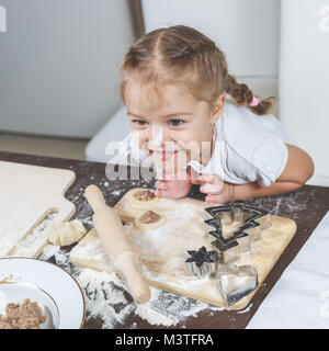 Funny little girl shows her tongue in kitchen preparing dough and dumplings Stock Photo