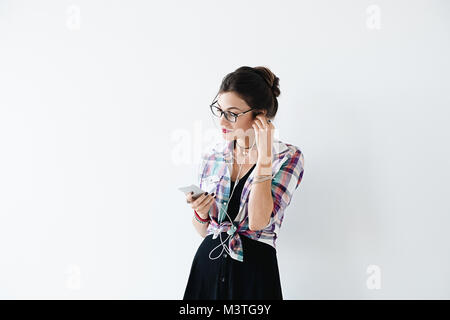 Stunning lady in plaid colorful shirt and glasses holds phone with one arm and put the headphones in the ears with smile, on a white background. Prett Stock Photo