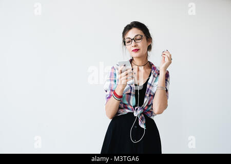 Stunning lady in plaid colorful shirt and glasses, is looking at phone screen and putting on her earphones with smile, on white background. Pretty gir Stock Photo
