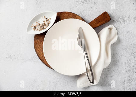 empty plate and cutlery on a wooden cutting board. a fork, a knife and a salt bowl with a pepper shaker. on white stone background, napkin. the table  Stock Photo
