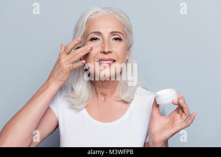 Perfect, pretty, woman applying eye cream, holding jar of cosmetic product looking at camera over gray background Stock Photo