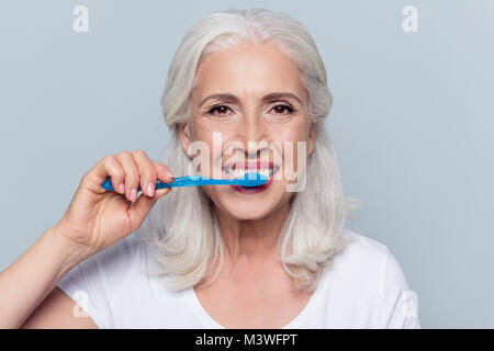 Concept of cleaning teeth is a correct way. Close up photo of happy cheerful mature old lady with good visage brushing her teeth with a blue toothbrus Stock Photo