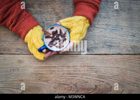 Womans hands with yellow woollen gloves holding a mug of hot chocolate with a cinnamon stick and dark chocolate shavings from above Stock Photo