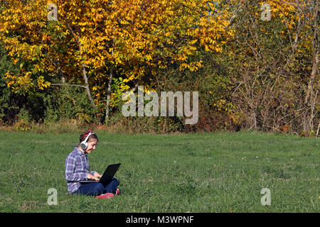 Happy little girl playing laptop and listening music on headphones in park Stock Photo