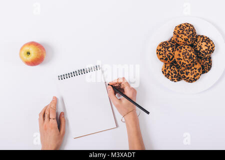 Women's hands write in a notebook, apple and cookies in a plate on a white table, top view Stock Photo