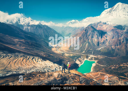 Standing men with backpacks on the mountain peak against beautiful mountain valley at sunset. Landscape with two man, rocks in clouds, blue sky, lake, Stock Photo