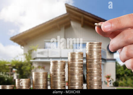 Human Hand Placing A Coin On Increasing Coin Stacks In Front Of House Stock Photo