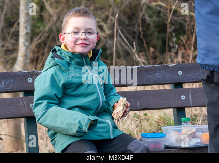 6 year old smiling boy wearing glasses sitting on a bench having a picnic lunch.  He is holding a ham and cheese roll, in Winter, Scotland, UK Stock Photo