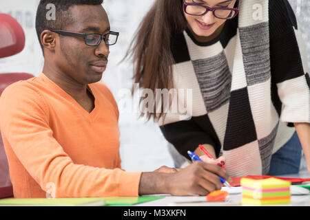 Two diverse work colleagues smiling and writing down notes while sitting together at a table in a modern office Stock Photo