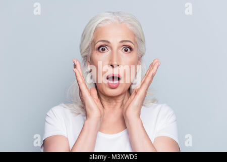 Pretty, nice, old, shocked, scared woman with wide open eyes and mouth in t-shirt holding palms near cheeks looking at camera over gray background Stock Photo