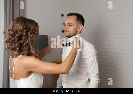 Bride fixes bow tie on groom s neck while they stand dressed in white shirt Stock Photo