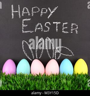 Happy Easter on chalkboard with row of Easter eggs in grass.Two with hand drawn Easter Bunny ears. Stock Photo