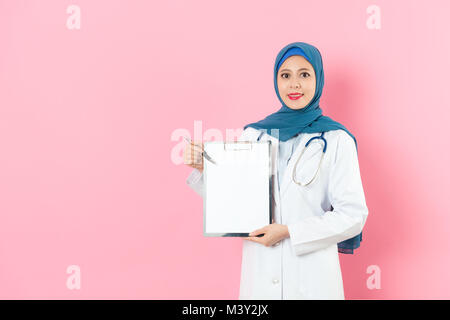 confident pretty muslim woman doctor holding clipboard looking at camera and using pen pointing white blank paper isolated on pink background. Stock Photo