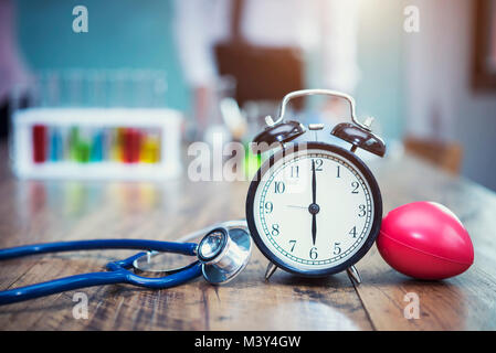 Healthy checking plan concept. Alarm clock with medical instruments stethoscope and red heart on wood table and blurred science lab accessories backgr Stock Photo