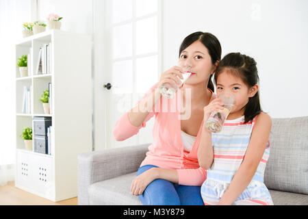 smiling young little daughter with mother sitting on sofa couch in living room together and drinking water looking at camera. Stock Photo