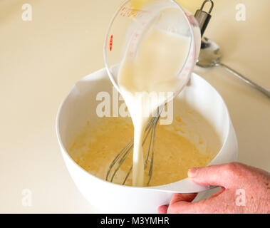 Man pouring milk into batter to make pancake batter for Shrove Tuesday with flour, milk, eggs, and oil in a home kitchen Stock Photo
