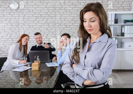 Portrait Of A Sad Businesswoman With Colleagues Laughing On Her In Background Stock Photo
