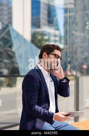 Smiling businessman sitting on a bench using a tablet and cellphone Stock Photo