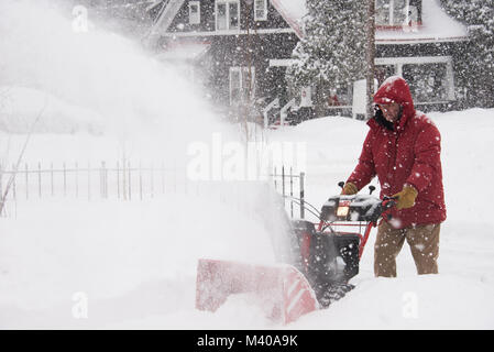 Man running a snowblower blowing snow from a driveway during a snowstorm in the Adirondack Mountains, NY USA Stock Photo