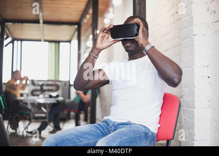 African young man wearing virtual reality glasses in modern interior design coworking studio. Smartphone using with VR goggles headset. Stock Photo