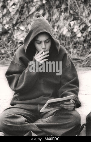 A teenage girl with a hoodie, sitting on a curb, smoking a cigarette, looking at a photo. Stock Photo