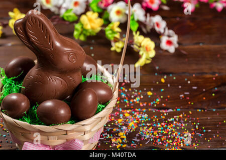 Beautiful Easter composition with chocolate bunny and eggs in a wicker basket with a bow, colored powder for cakes on old brown wooden background Stock Photo