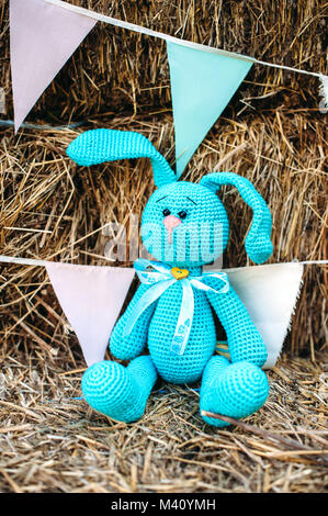 Knitted soft toy, blue rabbit on the straw. Handmade string of blue and pink flags on the background. Rustic style Stock Photo