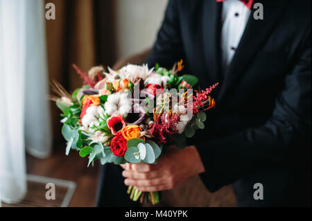 Groom holding wedding bouqet in his hands. Bouquet in boho, rustic style with cotton, purple callas, red roses. Closeup Stock Photo