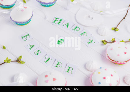 Holiday composition of Happy Easter lettering, branches with young shoots of greenery, decorated cupcakes, merengue sweets, bird figure on wooden back Stock Photo