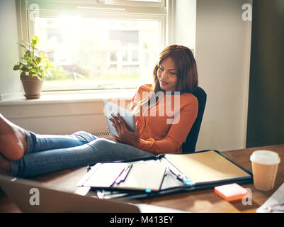 Smiling young African female entrepreneur working online with a digital tablet while leaning back in a chair in her home office Stock Photo