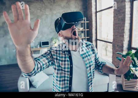 Bearded, shocked, wondered man in shirt with open mouth in motion, getting experience wearing vr goggles headset, watching video, trying to touch some Stock Photo