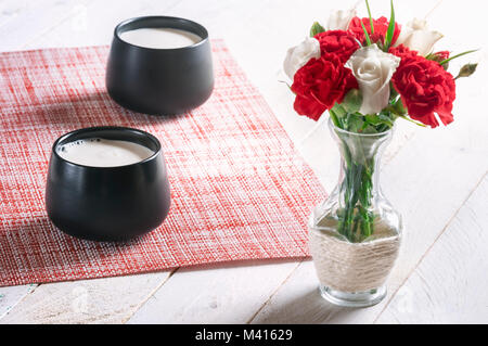 Breakfast table with two black mugs full of fresh milk and a bouquet of white roses and red carnations, on a white table, on a sunny morning. Stock Photo