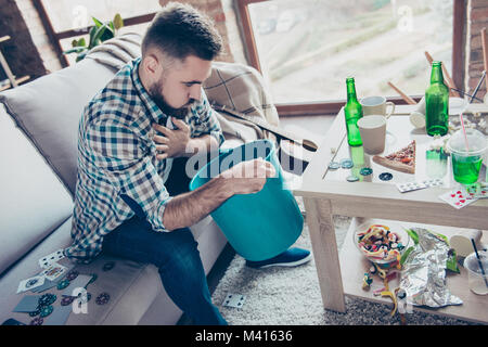 Bearded tired exhausted wearing casual checkered shirt is feeling unwell and suffering from nausea, he is holding a blue bucket Stock Photo