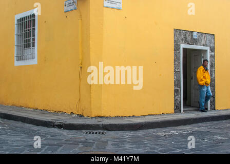 A man wearing jeans and a yellow jacket standing in the doorway or a colonial building painted yellow Stock Photo