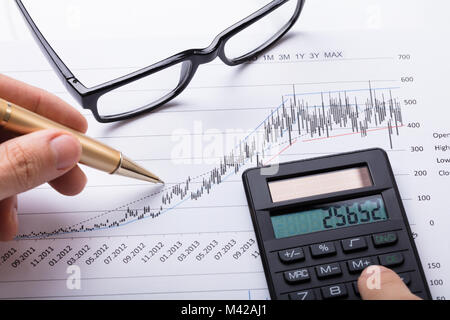Close-up Of Person Hand Using Calculator And Pen To Calculate Financial Chart On Table Stock Photo