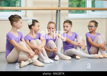 Young beautiful ballerinas talking in ballet studio. Group of little ballet dancers sitting on the floor and having a rest. School friends and childho Stock Photo
