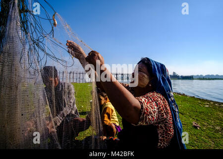 A fishermans woman is collecting fish from a net in front of U Bein Bridge, an old teakwood bridge, spanning over Taungthaman Lake Stock Photo