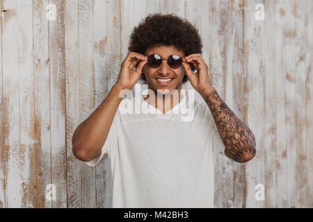 Choose a fashionable summer accessory! Portrait   man with stylish hairstyle and perfect smile adjusting his sunglasses. While standing against old wo Stock Photo