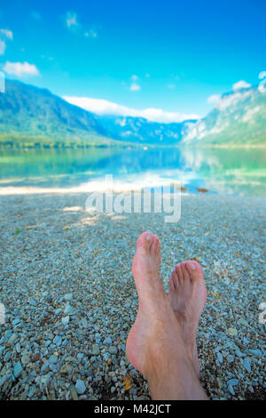 Summer holiday by the lake, barefoot adult caucasian male relaxing on beach pebbles and looking into water and mountains in the distance. Enjoying vac Stock Photo