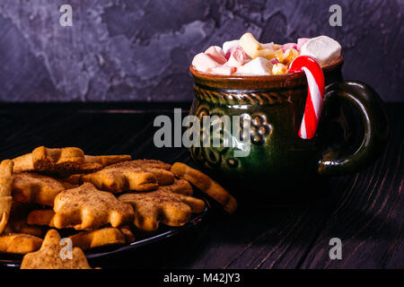 Ginger biscuits on a saucer next to a ceramic old cup full of marshmallocks and a New Year's candy in it against a background of dark concrete Stock Photo