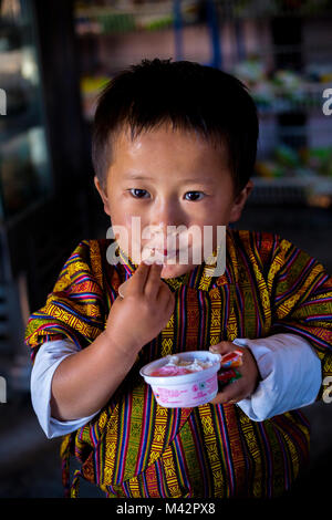 Punakha, Bhutan.  Young Boy in Traditional Male Gho Garment, Eating Ice Cream in the Lobeysa Market. Stock Photo