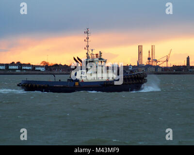 Harbor tug from the Port of Felixstowe makes its way back up the river against a background of orange sunset over seafront of Harwich, Suffolk, UK Stock Photo