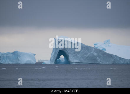 A large iceberg with an archway floating in Charlotte Bay in Antarctica. Dense clouds are overhead and steel gray water in the foreground. Stock Photo