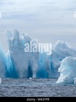 A light, medium and dark blue iceberg floating in the dark gray water of the Southern Ocean in Antarctic Sound. Overcast sky and a snow continent are  Stock Photo