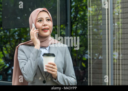 smiling professional female muslim business woman standing in outside relaxing and using mobile smartphone talking with friend chatting. Stock Photo