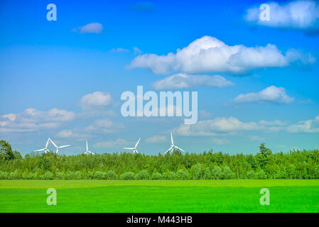 Summer scene of wind turbines behind forest, brightly green meadow, cloudy blue sky in evening, before sunset on countryside. Rural landscape Stock Photo