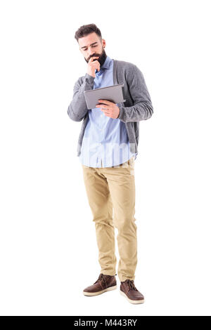 Doubtful bearded businessman with hand on chin looking at tablet computer. Full body length portrait isolated on white studio background. Stock Photo