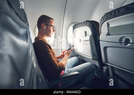 Connection in the airplane. Young man (traveler) using smart phone during flight and listening music. Stock Photo